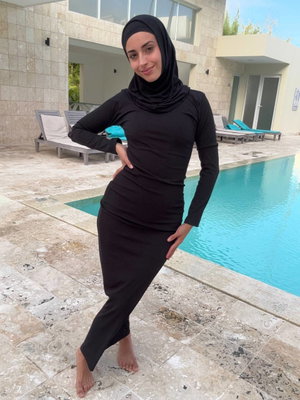 Woman in black hijab and long-sleeve dress posing by poolside, barefoot on stone tiles.