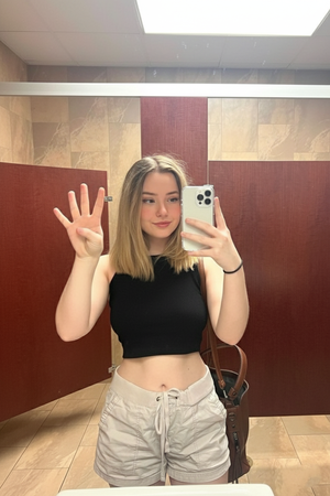 Woman with blonde hair in black crop top and beige shorts waving in mirror selfie.