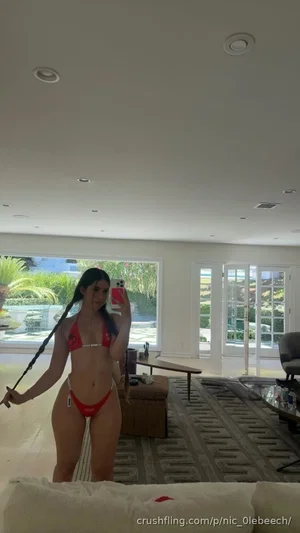 Woman in red bikini taking mirror selfie in bright living room with large windows.