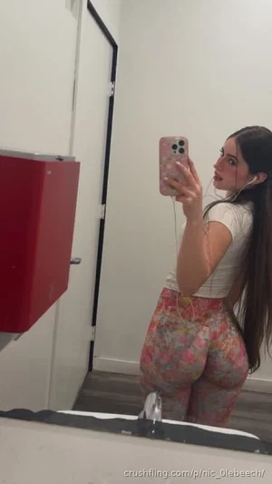 Woman with long dark hair in white crop top and floral pants taking mirror selfie in bathroom.