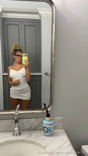 Woman in white dress taking mirror selfie in bathroom, blonde hair, glasses.