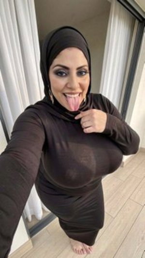 Woman in black hijab and sheer long-sleeve dress posing indoors, barefoot on wooden floor.