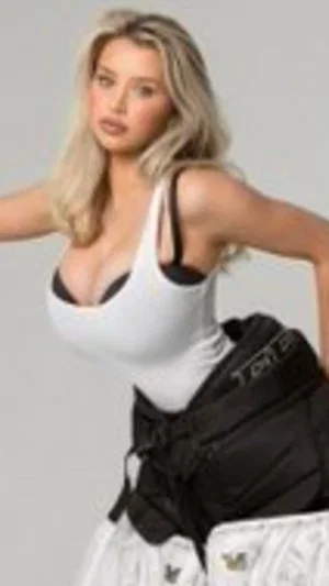 Blonde woman in white tank top and black harness, posing against gray background.