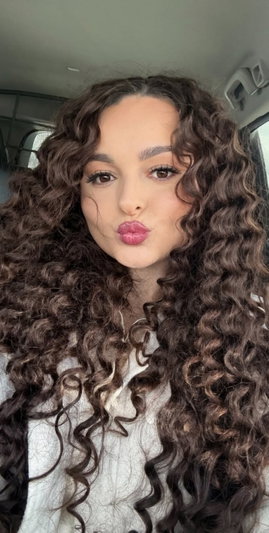 Woman with long curly brown hair in white top inside car.