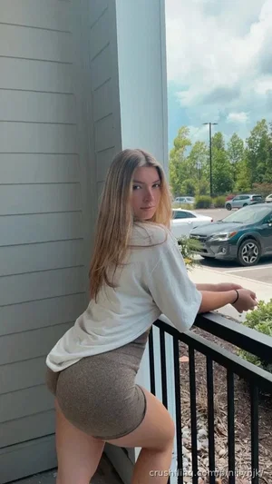 Woman with long hair in white shirt and brown shorts posing on balcony outdoors.