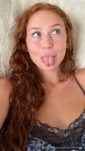 Woman with curly red hair in black lace top lying on textured surface, sticking out tongue.