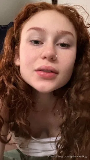Woman with curly red hair wearing a white top, indoors, close-up selfie.
