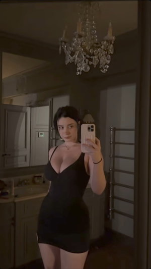 Woman in black dress taking mirror selfie in bathroom with chandelier.