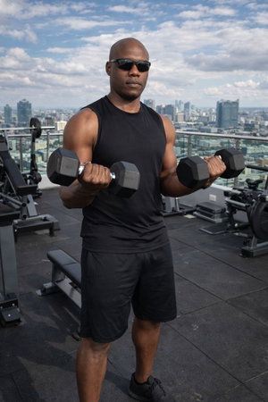 Athletic bald Black man in tank top and shorts lifting dumbbells on rooftop gym.