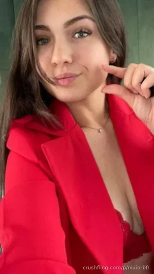 Woman in red blazer and red lingerie, posing indoors with hand near face.