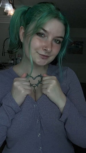 Woman with green pigtails in purple cardigan showing chest tattoo.