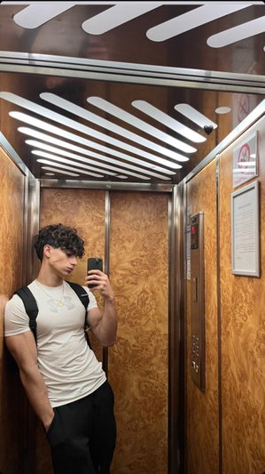 Man with curly hair in white t-shirt and black pants taking mirror selfie in elevator.