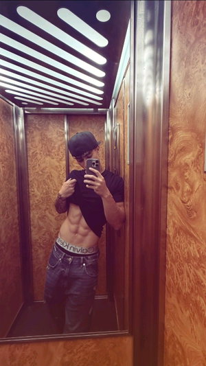 Man in black t-shirt and jeans takes mirror selfie in elevator, showing abs.