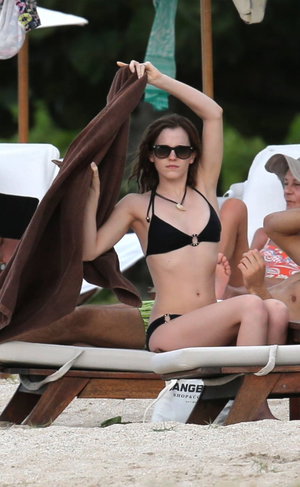 Woman in black bikini sitting on beach lounge, holding towel, wearing sunglasses.