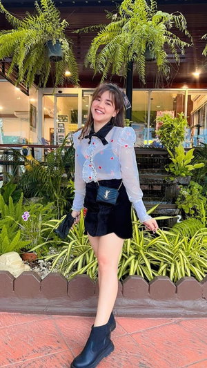 Woman in sheer blouse and black skirt standing outdoors among plants.