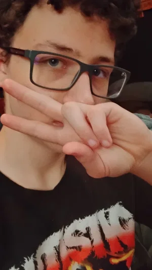Person with curly hair and glasses making a hand gesture, wearing a black graphic t-shirt indoors.