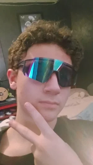 Young man with curly hair wearing reflective sunglasses, making a peace sign indoors.