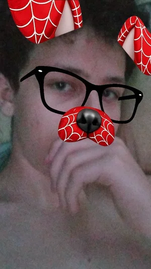 Shirtless man with curly hair using a Spider-Man dog filter, hand near mouth, indoor setting.
