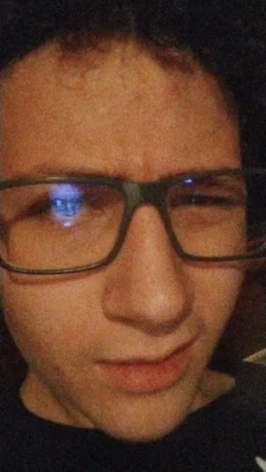Close-up of a person with curly hair and glasses, wearing a dark shirt indoors.