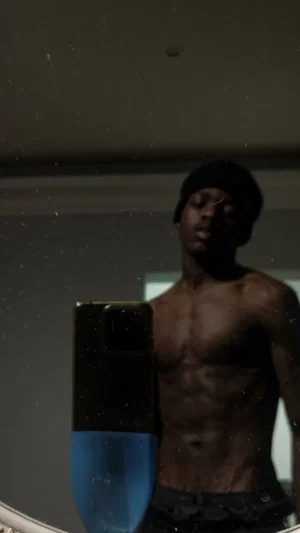 Shirtless man with defined muscles wearing dark pants and beanie, taking mirror selfie in dimly lit