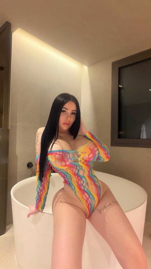 Curvy woman in sheer colorful mesh bodysuit by bathtub in bathroom.