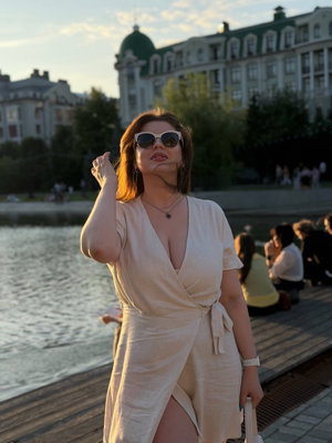 Woman in beige wrap dress with deep V-neck, standing by waterfront, wearing sunglasses.