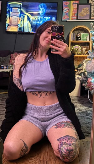 Tattooed woman in purple outfit taking mirror selfie indoors.