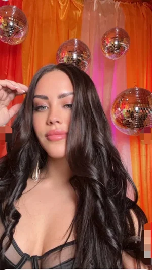 Woman with long dark hair in black sheer top, posing in front of orange curtains with disco balls.