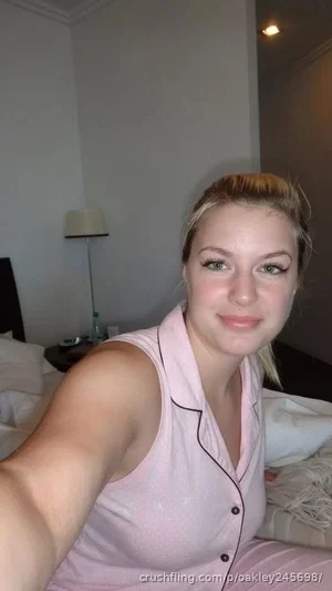 Woman in pink pajamas taking selfie in bedroom, blonde hair tied up.