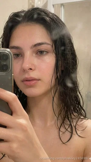 Woman with wet dark hair taking mirror selfie, bare shoulders, indoor bathroom setting.