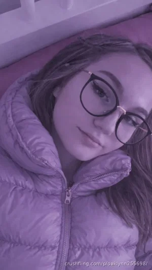 Woman with glasses lying on pink pillow, wearing purple puffer jacket, indoor setting.