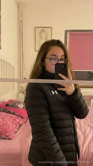Woman in black puffer jacket takes mirror selfie in bedroom with pink bedding.
