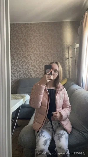 Woman in pink puffer jacket and tie-dye pants sits on couch, taking mirror selfie indoors.