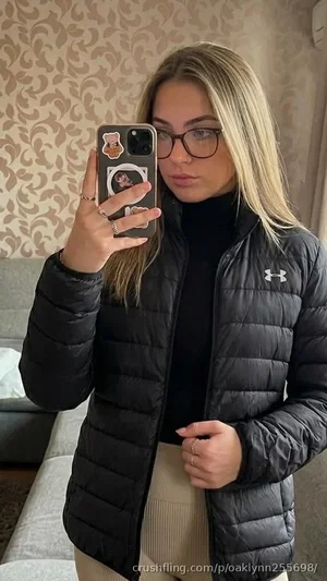 Woman with blonde hair and glasses wears black puffer jacket and beige skirt, taking mirror selfie