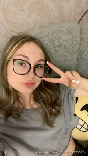 Woman with glasses and wavy hair in gray top, making peace sign on couch.