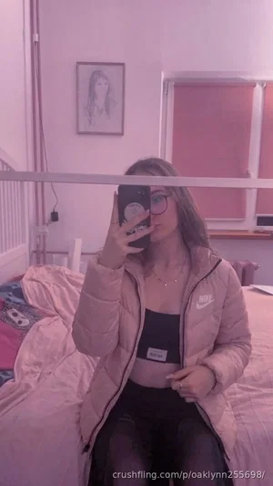 Woman in pink puffer jacket and black crop top sits on bed, taking mirror selfie in bedroom.