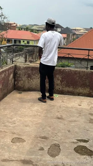 Man in white t-shirt and black pants standing on rooftop, facing away, overlooking residential area.