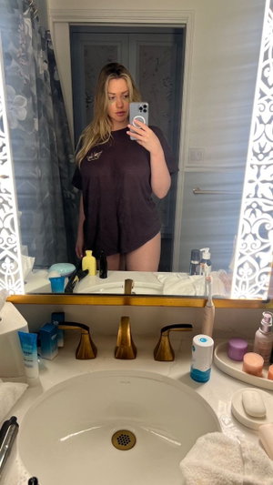 Woman with blonde hair taking mirror selfie in bathroom wearing t-shirt.