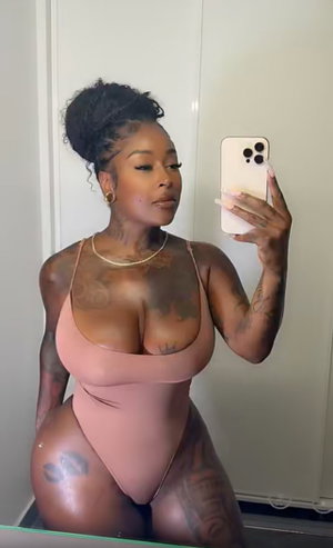Tattooed woman in pink bodysuit taking mirror selfie indoors.
