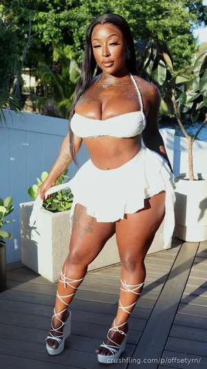 Woman in white bikini top and skirt, standing outdoors on deck with plants.