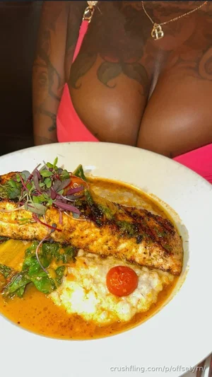 Woman with tattoos in pink top, holding plate of grilled fish and rice.