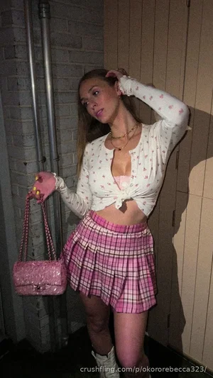 Woman in white floral top and pink plaid skirt, holding pink bag, standing indoors near brick wall.