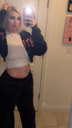Woman with blonde hair in white crop top and black jacket, standing indoors taking mirror selfie.