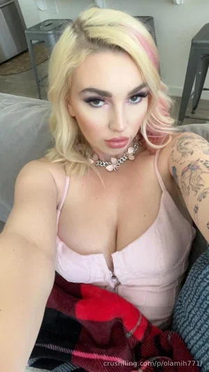 Woman with blonde hair and tattoos wears pink top, sitting on couch indoors.