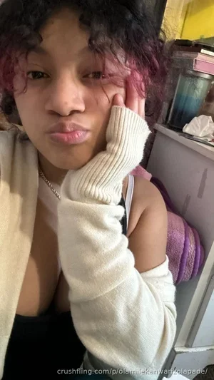 Woman with curly hair and pink highlights wearing a white sweater and black top, posing indoors.