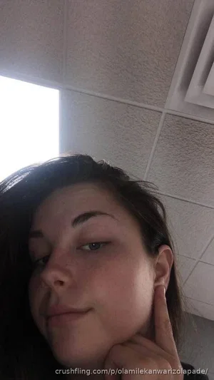 Woman with dark hair posing indoors, hand near face, ceiling tiles in background.
