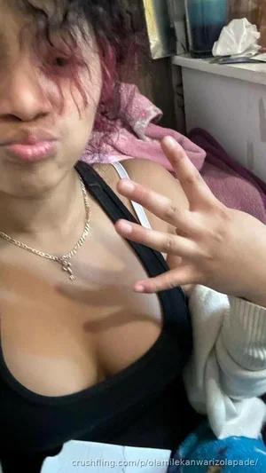 Woman with curly hair in black top and gold necklace, making hand gesture indoors.