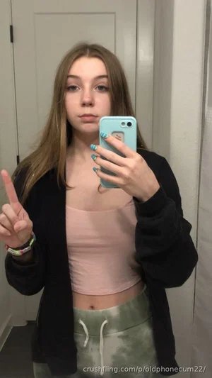 Woman with long hair in pink crop top and green tie-dye pants, taking mirror selfie indoors.
