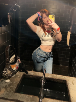 Red-haired woman in crop top and jeans taking bathroom selfie.
