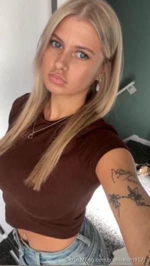 Blonde woman in brown crop top and jeans, tattoo on arm, indoor setting.
